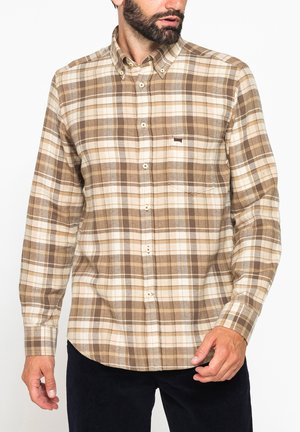 Uomo che indossa una camicia a quadri marrone e beige con bottoni e tasca frontale, in piedi davanti a uno sfondo bianco.