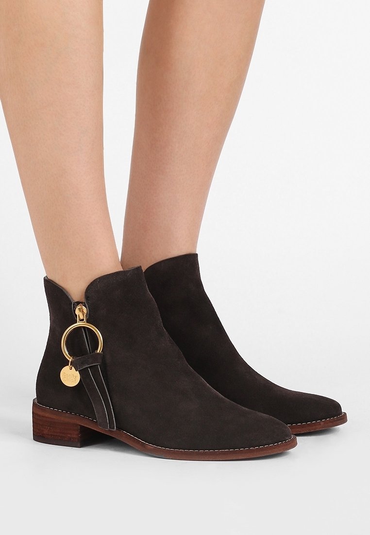 Mörkbruna mocka ankelskor med spetsig tå, guldringdetaljer, dragkedjedetaljer och en träklack, med synliga sömnadsdetaljer.