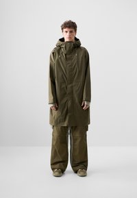 Olivegrüne wasserdichte Jacke mit Kapuze, lockere Passform, langen Ärmeln und Frontreißverschluss; kombiniert mit lockeren olivfarbenen Hosen und beigen Schuhen.