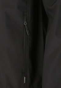 Musta takki, jossa on vetoketjullinen tasku, sileä pinta ja huomaamaton brändäys vasemmassa alakulmassa. Suunnittelu on toiminnallinen ja minimalistinen.