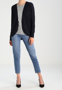 Laivastonsininen avokardigan harmaan paidan päällä, yhdistettynä vaaleansisiin nilkkapituisiin farkkuihin ja mustiin korkokenkiin; sileä kangas ja rento istuvuus.