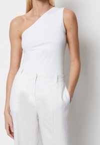 Nainen, jolla on päällään valkoinen olkaimeton toppi, joka on tussahtunut valkoisiin korkeavyötäröisiin housuihin, yksi käsi taskussa.