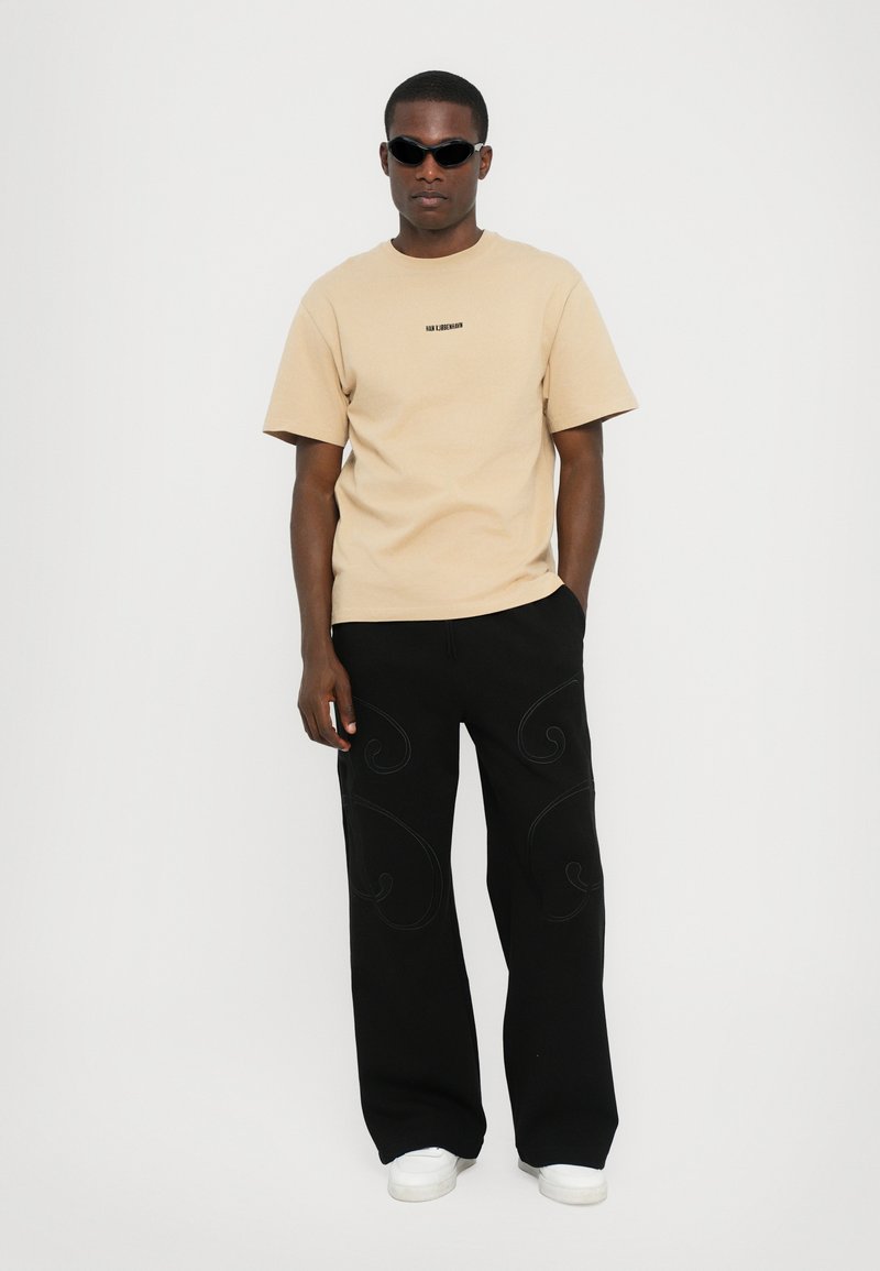 T-shirt en coton beige à manches courtes avec logo sur la poitrine, associé à un pantalon large noir orné de motifs en broderie tourbillonnants et des chaussures blanches.