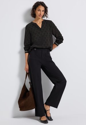 Schwarz gemusterte Bluse mit langen Ärmeln; schwarze weite Hose; braune Wildleder-Handtasche; schwarze Schuhe mit Knöchelriemen.