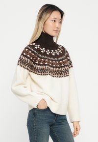 Sweater de malha em creme com um colarinho alto castanho escuro. Apresenta um padrão geométrico em tons de castanho e branco nos ombros e nas mangas.