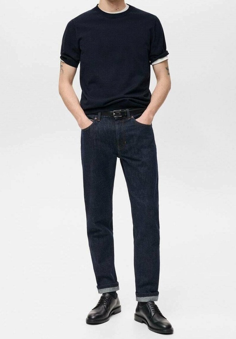 Homme portant un t-shirt noir aux manches retroussées, un jean bleu foncé retroussé, une ceinture noire et des chaussures en cuir noires, debout les mains dans les poches.