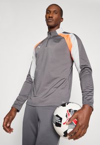 Šedá sportovní bunda s oranžovými a bílými akcenty. Obsahuje zipový límec, dlouhé rukávy a je kombinována se šedými kalhotami a černo-bílým fotbalovým míčem.