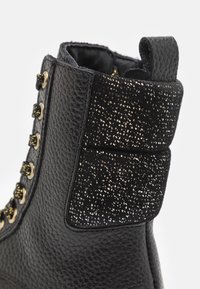 Černá kožená bota s texturovaným svrškem, lesklým zlatým kováním, třpytivým černým podpatkem a poutkem pro snadné obouvání.