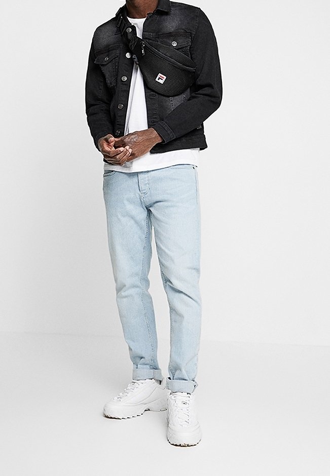 Uomo che indossa una giacca di denim nera, una t-shirt bianca, dei jeans azzurri arrotolati, delle sneakers bianche e una borsa a tracolla nera, in piedi contro uno sfondo bianco.