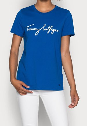 Osoba ubrana w niebieską koszulę z krótkim rękawem z białym napisem „Tommy Hilfiger” oraz białe spodnie, stojąca na jednolitym tle.
