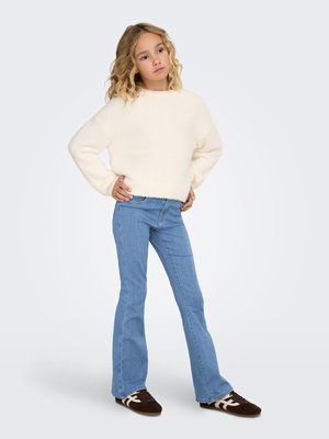 White knitted sweater with wide sleeves, paired with light blue flared jeans and brown sneakers with white accents. Hair is wavy.
