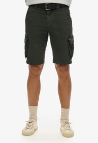 Shorts cargo jusqu'aux genoux en vert olive, avec plusieurs poches latérales, un devant plat et un tissu texturé. Assortis avec des baskets légères et des chaussettes.