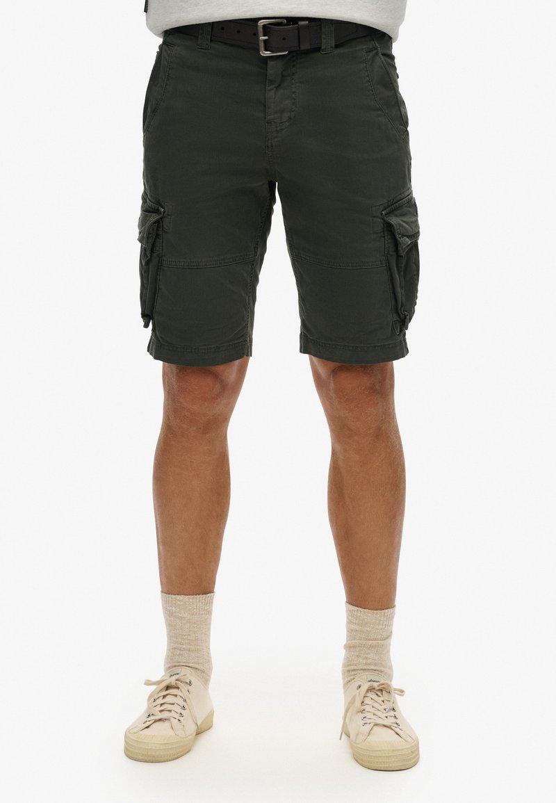 Shorts cargo jusqu'aux genoux en vert olive, avec plusieurs poches latérales, un devant plat et un tissu texturé. Assortis avec des baskets légères et des chaussettes.