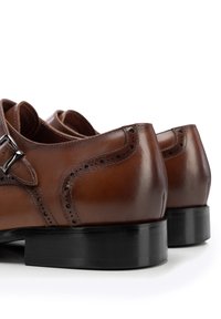 Zapatos de cuero marrón con un acabado pulido, que presentan detalles de brogue, una hebilla plateada y un talón negro sólido.