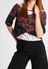 Blusa con motivo floreale che presenta uno sfondo nero con accenti rossi e bianchi, maniche corte e un design a strati su una base bianca.