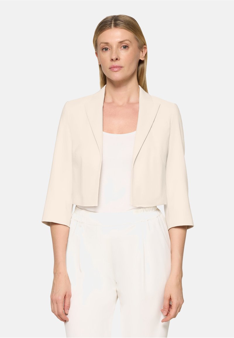 Femme portant un blazer court beige clair à manches 3/4 et un pantalon blanc taille haute, debout devant un fond clair uni.