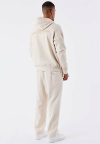 Beige hoodie met een groot logo op de achterkant, gecombineerd met bijpassende loose-fit broek en witte sneakers. Gestructureerde stof, casual uitstraling.