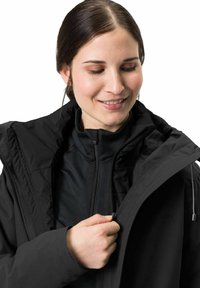 Schwarze isolierte Jacke mit einem Reißverschluss und hohem Kragen. Verfügt über eine Kapuze und strukturstoff. Trägt man über einem schwarzen Oberteil.