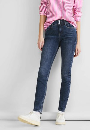 Jeans skinny en denim bleu foncé, arborant une taille moyenne, un design à cinq poches et des coutures contrastées. Assortis à des baskets blanches.