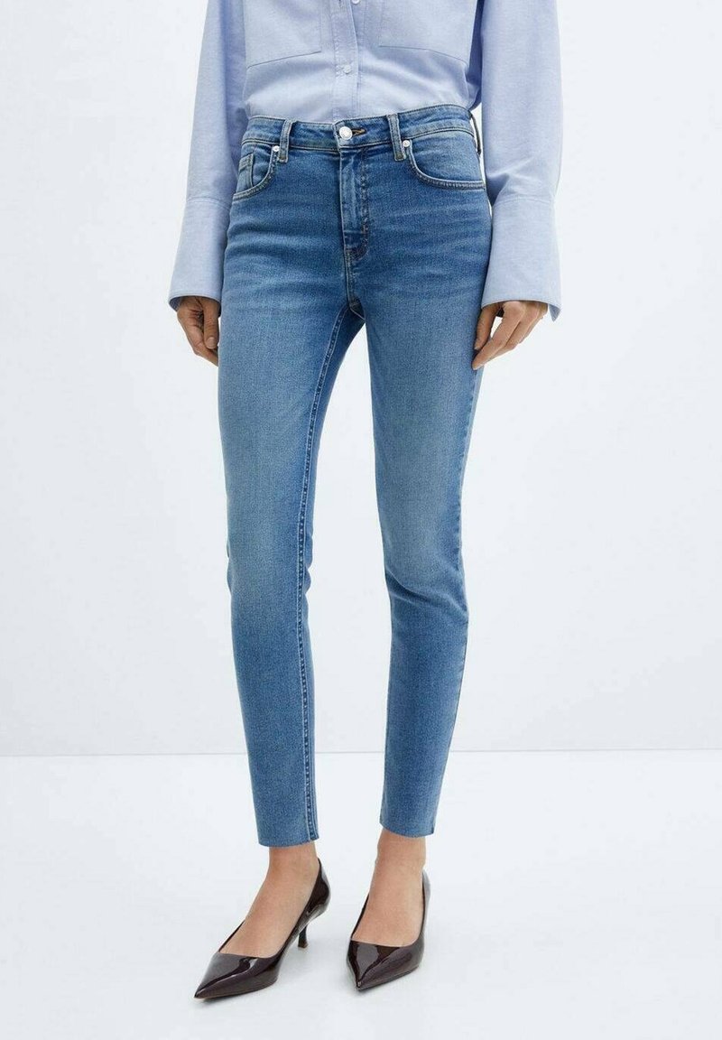 Femme portant un jean bleu clair, une chemise boutonnée bleu clair et des escarpins à bouts pointus marron foncé, debout sur un sol blanc.