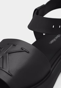 Μαύρο σανδάλι ανοιχτής μύτης με ανάγλυφο μεγάλο λογότυπο "CK" στο μπροστινό λουρί και ρυθμιζόμενο λουρί στον αστράγαλο με αγκράφα.