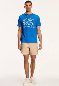 Niebieska bawełniana koszulka z białym graficznym napisem „HOLIDAY OFTEN”. W zestawie beżowe szorty typu cargo i białe trampki.