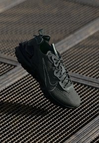 Scarpa sportiva verde scuro realizzata in materiale sintetico con un design testurizzato, lacci davanti e suola arrotondata. Presenta un dettaglio del logo ben visibile.