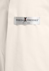 Biała tkanina z naszytą metką, na której znajduje się napis "FRIEDA FREDDIES NEW YORK", z małym czarnym emblematem psa i czerwonym detalem kołnierza.