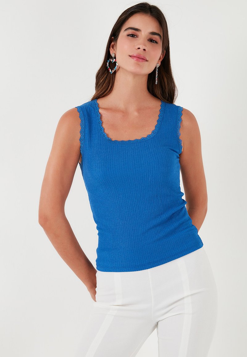 Top tank blu ribbed con scollatura quadrata e orlo scallop. Indossato con pantaloni bianchi a vita alta, che mettono in evidenza una silhouette aderente.