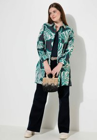 Camicia a maniche lunghe con un motivo a foglie verdi e bianche, abbinata a pantaloni larghi scuri. Tiene in mano una piccola borsa intrecciata nera e beige.