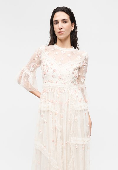 Γυναίκα με σκούρα μαλλιά που φορά ένα διάφανο λευκό φόρεμα διακοσμημένο με ροζ και λευκή κεντητική με λουλούδια και λεπτομέρειες με βολάν, στέκεται μπροστά σε απλό φόντο.