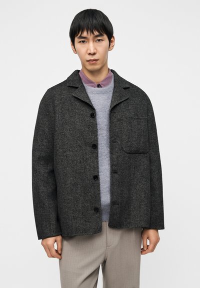 Σκούρο γκρι σακάκι με σχέδιο καρό ψαροκόκαλο και άνετη εφαρμογή, με γιακά, μπροστινή τσέπη και μαύρα κουμπιά, φορεμένο πάνω από ανοιχτό γκρι πουλόβερ.