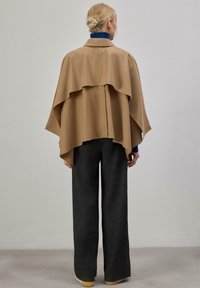 Personne aux cheveux blonds attachés en chignon, portant une cape beige, un col roulé bleu, un pantalon large gris foncé et des chaussures blanches, debout contre un mur uni.