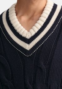 Námořnicky modrý svetr s vrabčím vzorem a výstřihem do V, s tlustým krémovým žebrovaným límcem a bílými šitími zdobícími linii výstřihu.