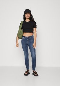 Sort cropped t-shirt, blå skinny jeans, sorte sandaler og grøn, kviltet skuldertaske. Model med langt, bølget hår og kasket.