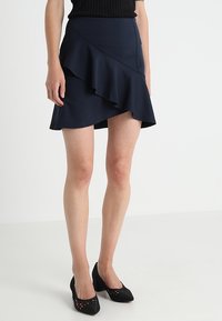 Femme portant une jupe asymétrique volantée bleu marine avec des chaussures à talons moyens en tricot noirs, se tenant devant un fond clair.