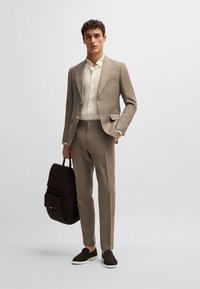 Light brown linen suit with a fitted silhouette. Paired with a cream button-up shirt and dark brown slip-on shoes. Holds a dark backpack.