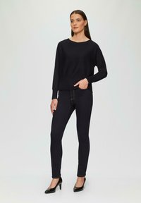 Pull noir avec ourlet côtelé, associé à un jean skinny noir. Le modèle porte des chaussures à talons noirs, se tenant devant un fond neutre.