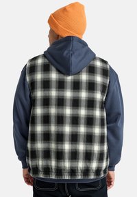 Black and white plaid vest with a soft texture, layered over a blue hoodie. The outfit is completed with an orange knit beanie.