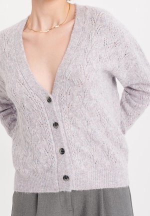Donna che indossa un cardigan lavorato a trecce grigio chiaro abbottonato sul davanti, abbinato a pantaloni grigi larghi e una sottile collana d'oro.
