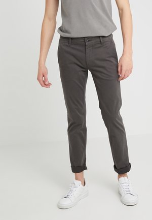 Persoon met opgerolde donkergrijze broek, witte sneakers en een lichtgrijs t-shirt dat in de broek is gestopt, staand tegen een eenvoudige lichte achtergrond.