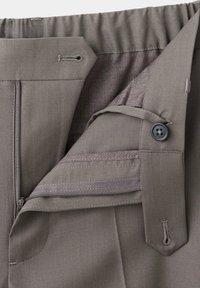Gros plan sur la ceinture d'un pantalon gris montrant un bouton, des boutonnières, une fermeture éclair et un dos élastiqué pour un ajustement réglable.