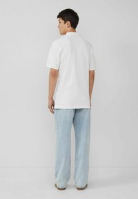 Weißes Poloshirt mit kurzen Ärmeln, klassischem Kragen und geradem Saum, kombiniert mit hellblauen, weit geschnittenen Jeans und hellbraunen Schuhen.