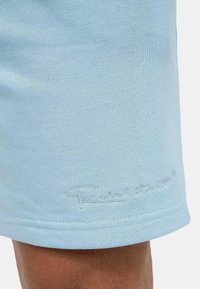 Shorts bleu clair en tissu texturé avec un logo brodé en fil blanc près de l'ourlet, présentant des lignes épurées et un design minimaliste.