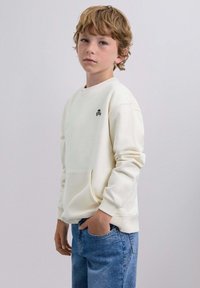 Crèmekleurig sweatshirt met een ronde hals, lange mouwen en een voorzak; heeft een klein zwart schedel logo op de borst. Draagt met blauwe jeans.