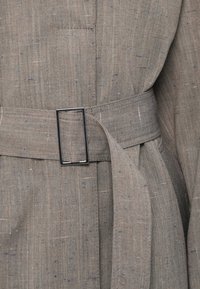 Γκρι λινάτσα ζώνη με ορθογώνια μεταλλική αγκράφα. Διαθέτει υφή υφάσματος με διακριτικές αποχρώσεις χρώματος. Περιλαμβάνει πλαϊνή τσέπη στο ένδυμα.