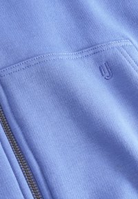 Felpa azzurra con zip, dal texture morbido. Presenta un piccolo logo e cuciture a contrasto. Primo piano dei dettagli del tessuto.