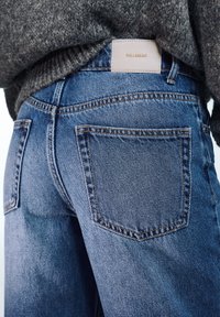 Halványkék színű, egyenes szabású farmernadrág. Jellemzői közé tartozik egy bőr címke "PULL&BEAR" felirattal és kontrasztos narancssárga varrás.