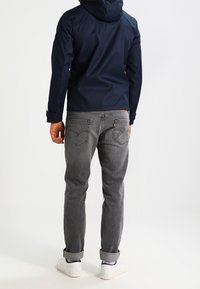 Mörkblå huvjacka, grå jeans med uppvikta fållar och vita sneakers. Förstärkta sömmar på fickorna, slitstarkt tyg.