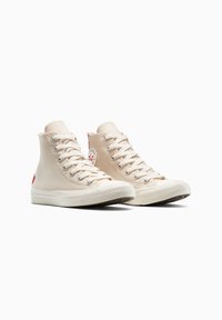Zapatillas de lona altas en color beige con suelas de goma blanca, con ojales de metal, cordones y un parche con el logo rojo en el lateral.